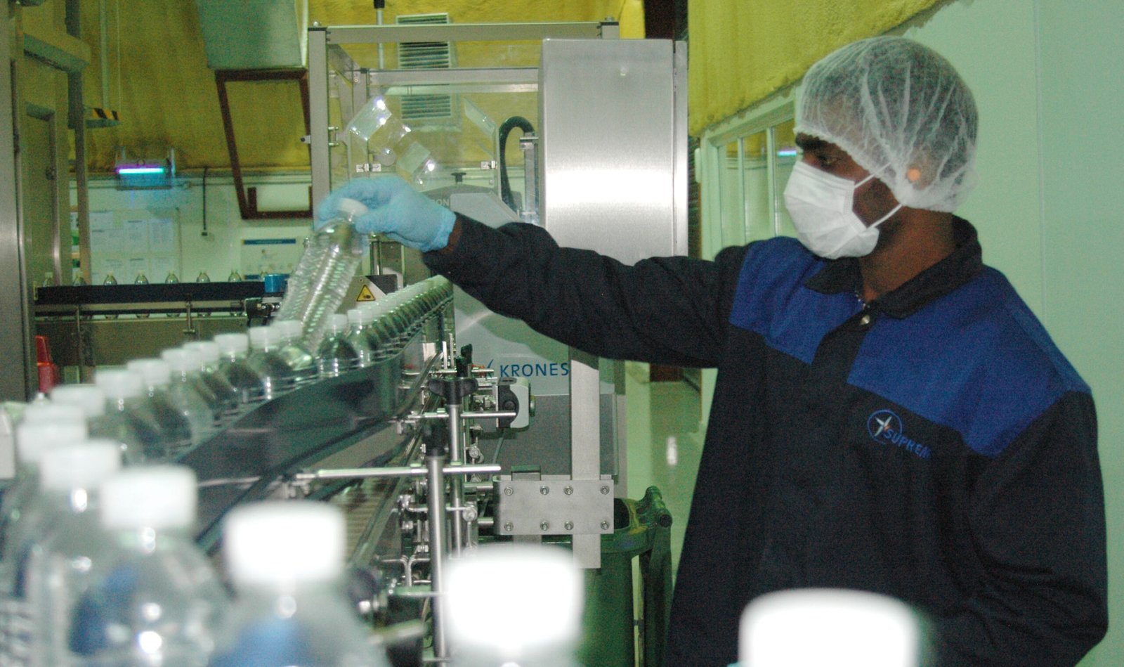 Worker in protective gear inspecting bottled water on a conveyor line during quality control at a bottling plant